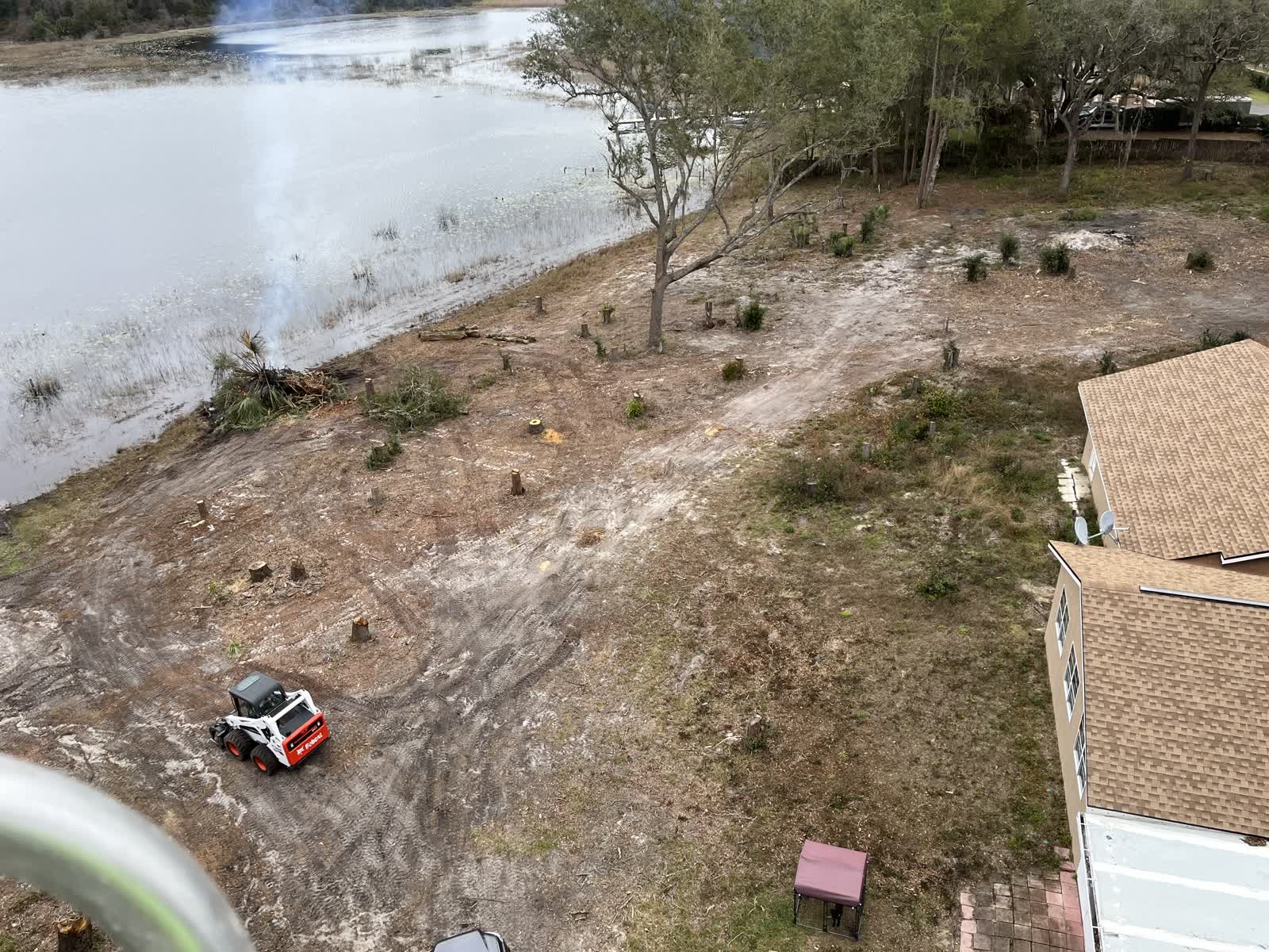 Aerial drone view of a cleared lot beside a Florida pond, with a skid steer working and a controlled burn pile smoking at the tree line