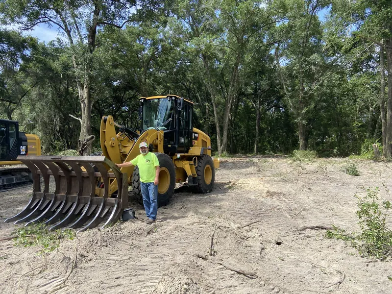 Owner standing beside a CAT wheel loader with a brush rake attachment on a freshly cleared parcel of land