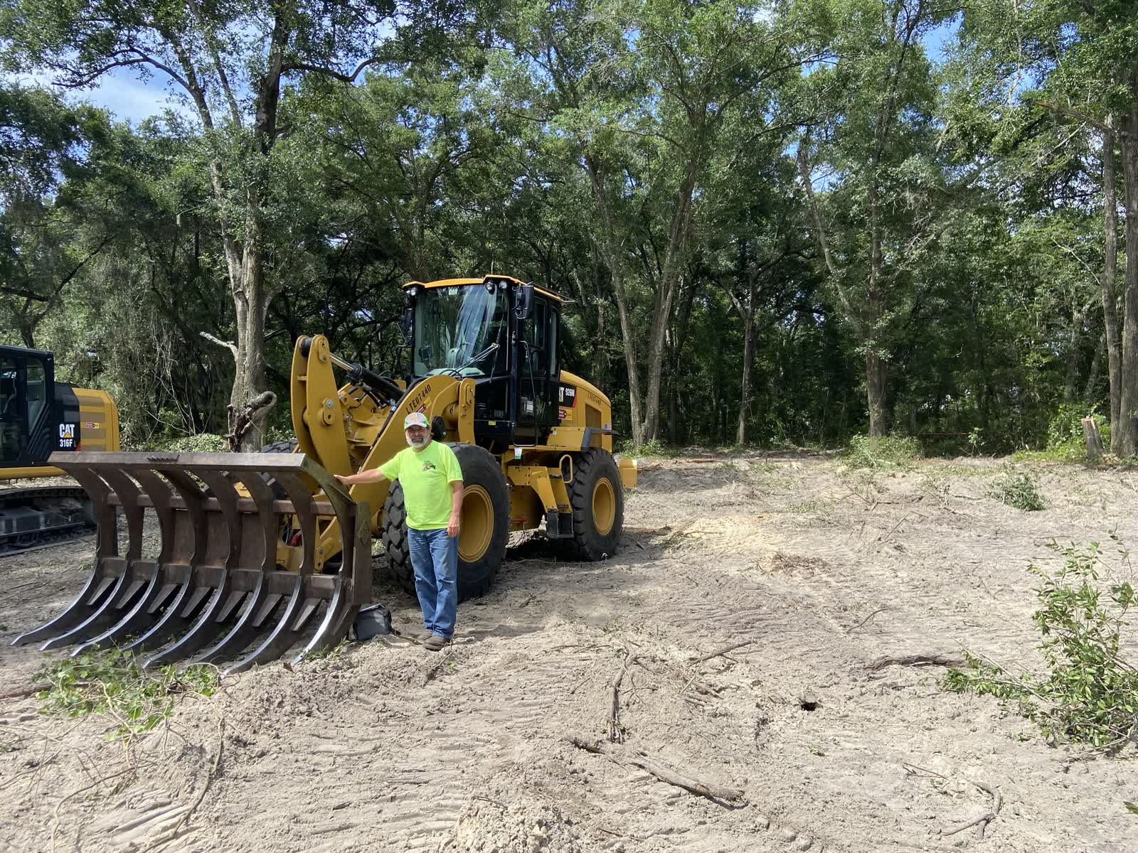 Owner standing beside a CAT wheel loader with a brush rake attachment on a freshly cleared parcel of land